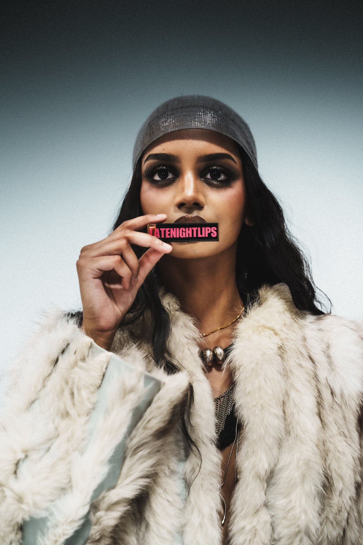 Model in a metallic headscarf and faux-fur coat holding a LATENIGHTLIPS gloss tube to her lips