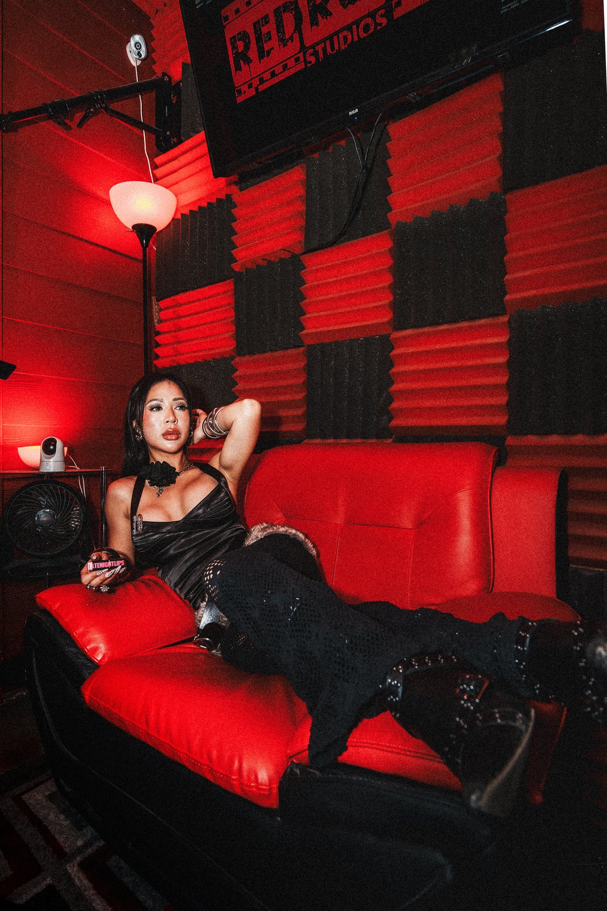 Model reclining on a red couch in a black outfit under red lighting