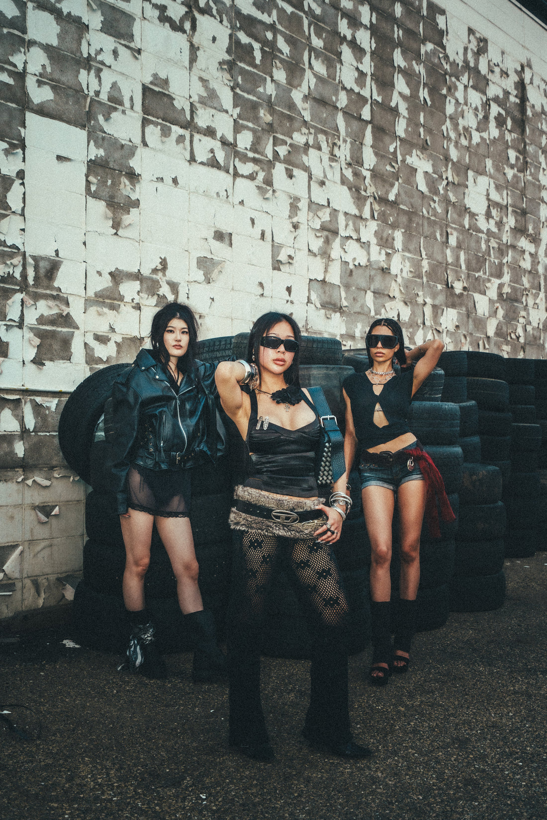 Three models in dark outfits posing beside stacked tires against a weathered industrial wall