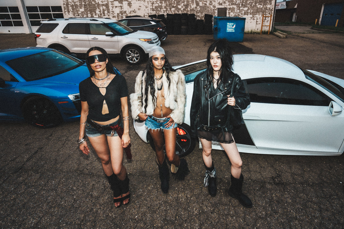Three LATENIGHTLIPS models posing between cars at an industrial lot.