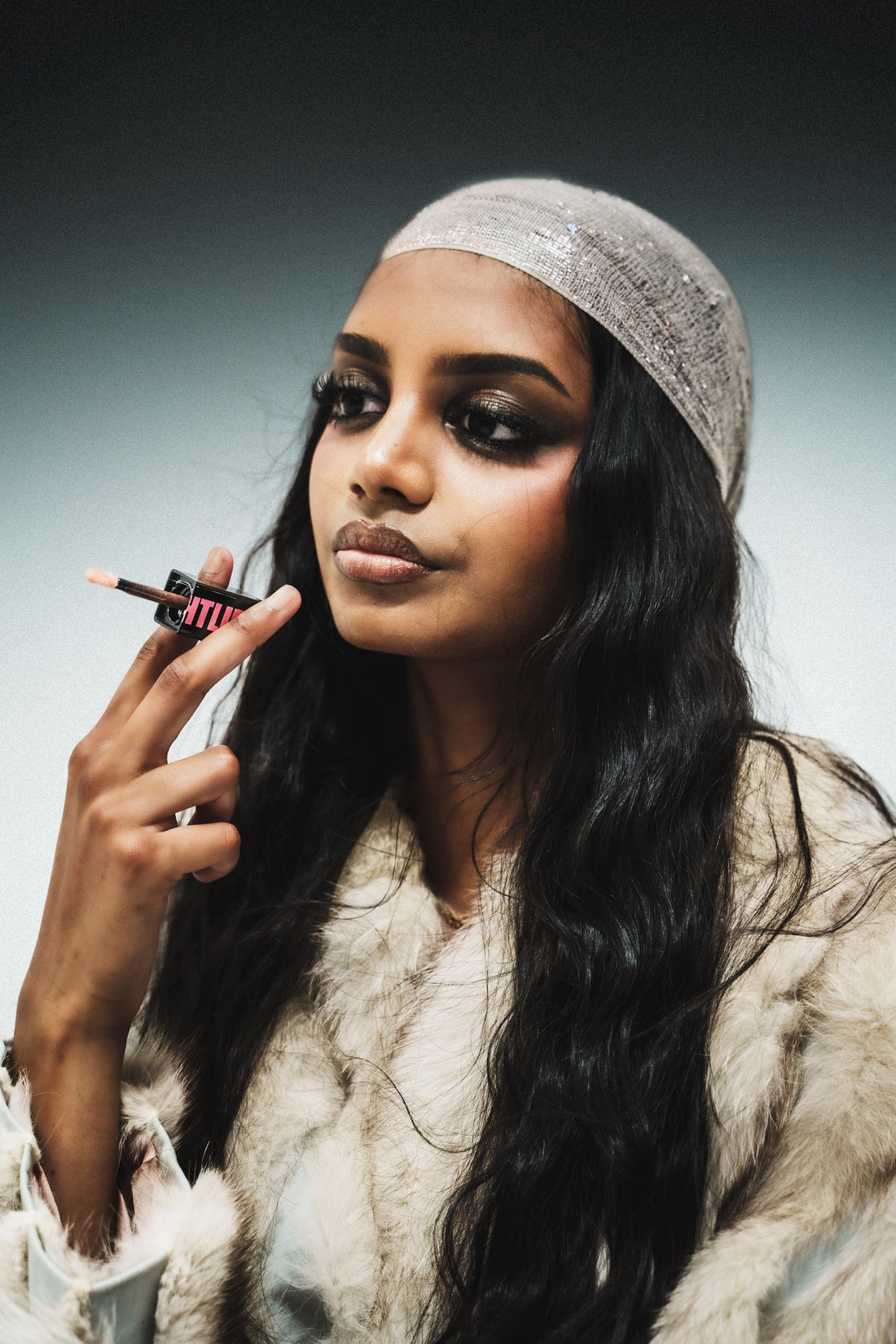 Model in a white headscarf and cream faux-fur coat applying gloss over a coloured lip