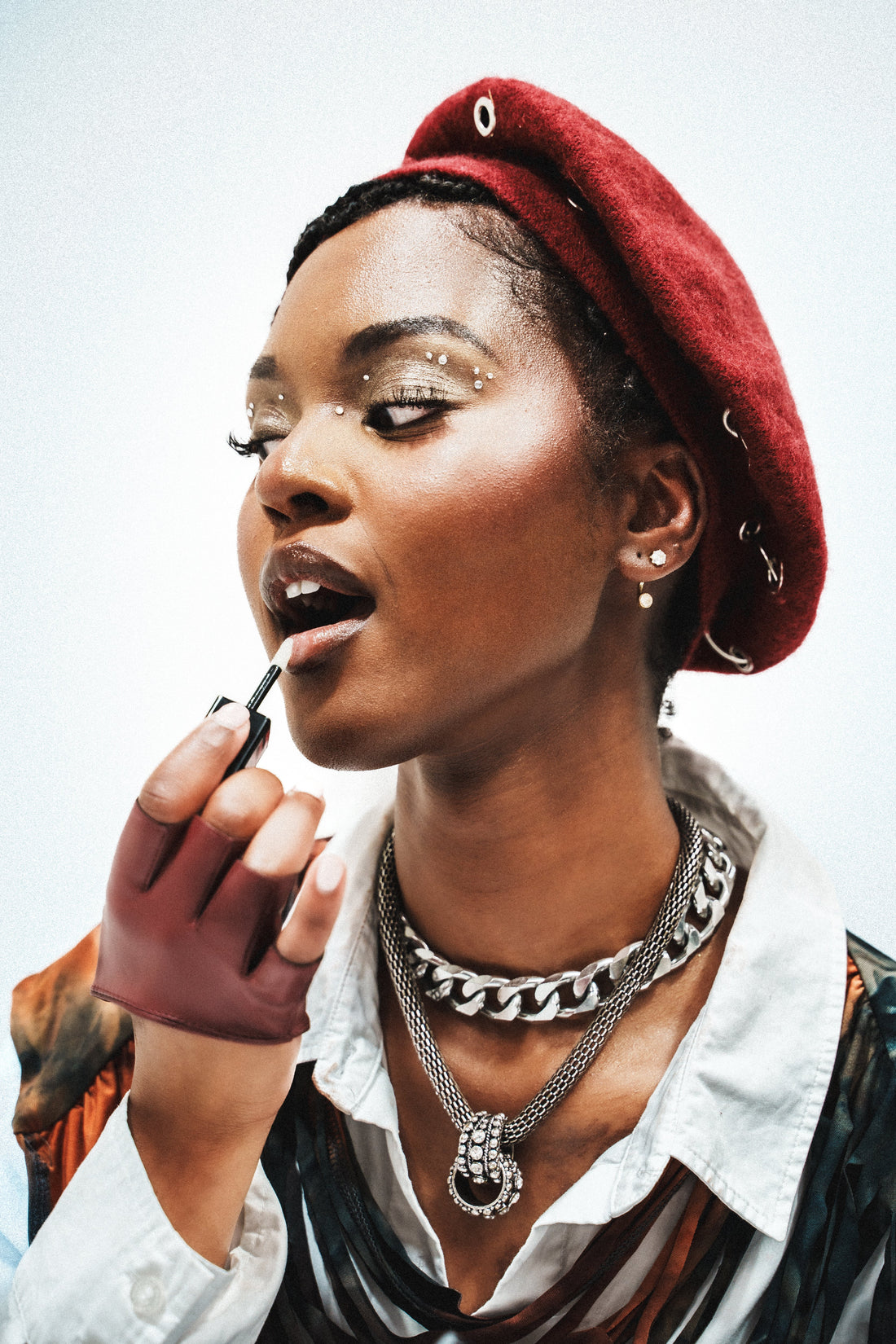 Model in a red beret applying lip gloss with a wand, wearing layered silver necklaces