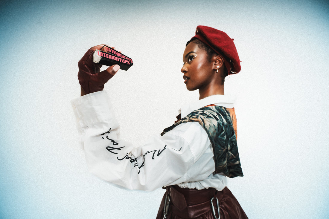 Model in profile wearing a red beret and patterned vest, holding black LATENIGHTLIPS packaging up against a pale studio background