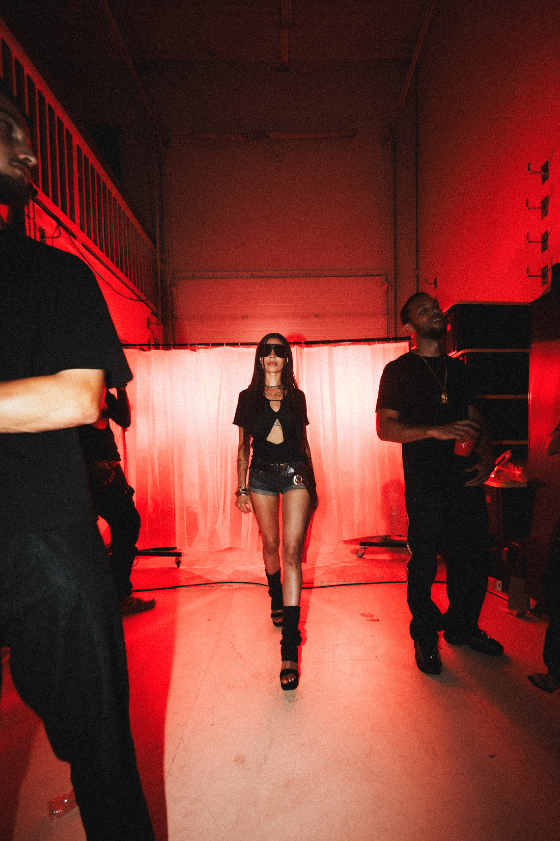 Woman in dark sunglasses and a black cutout outfit walking through a red-lit backstage room