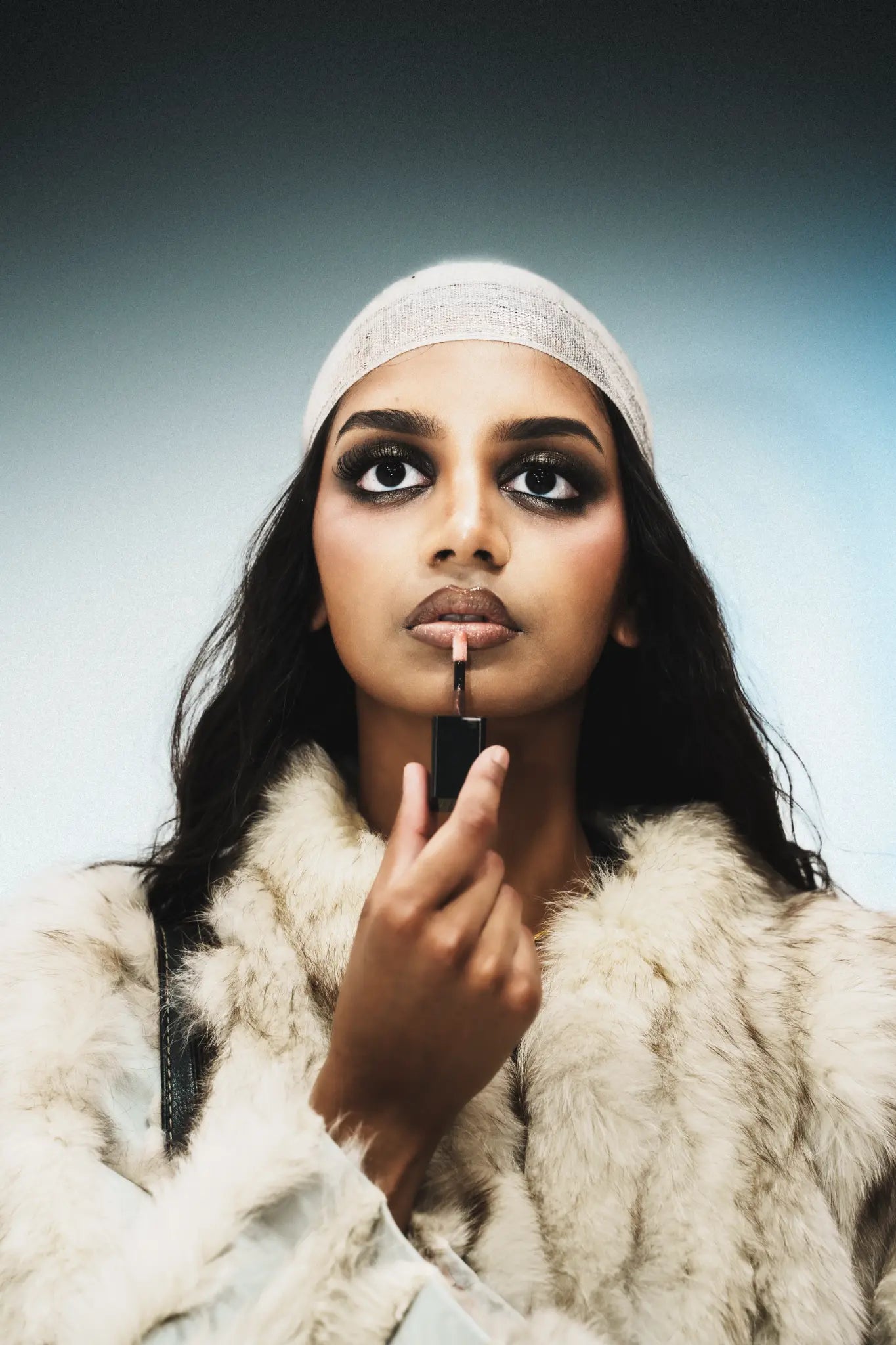 Celine holding Butter'd Up Lip Glaze close to her lips in a headscarf and fur.